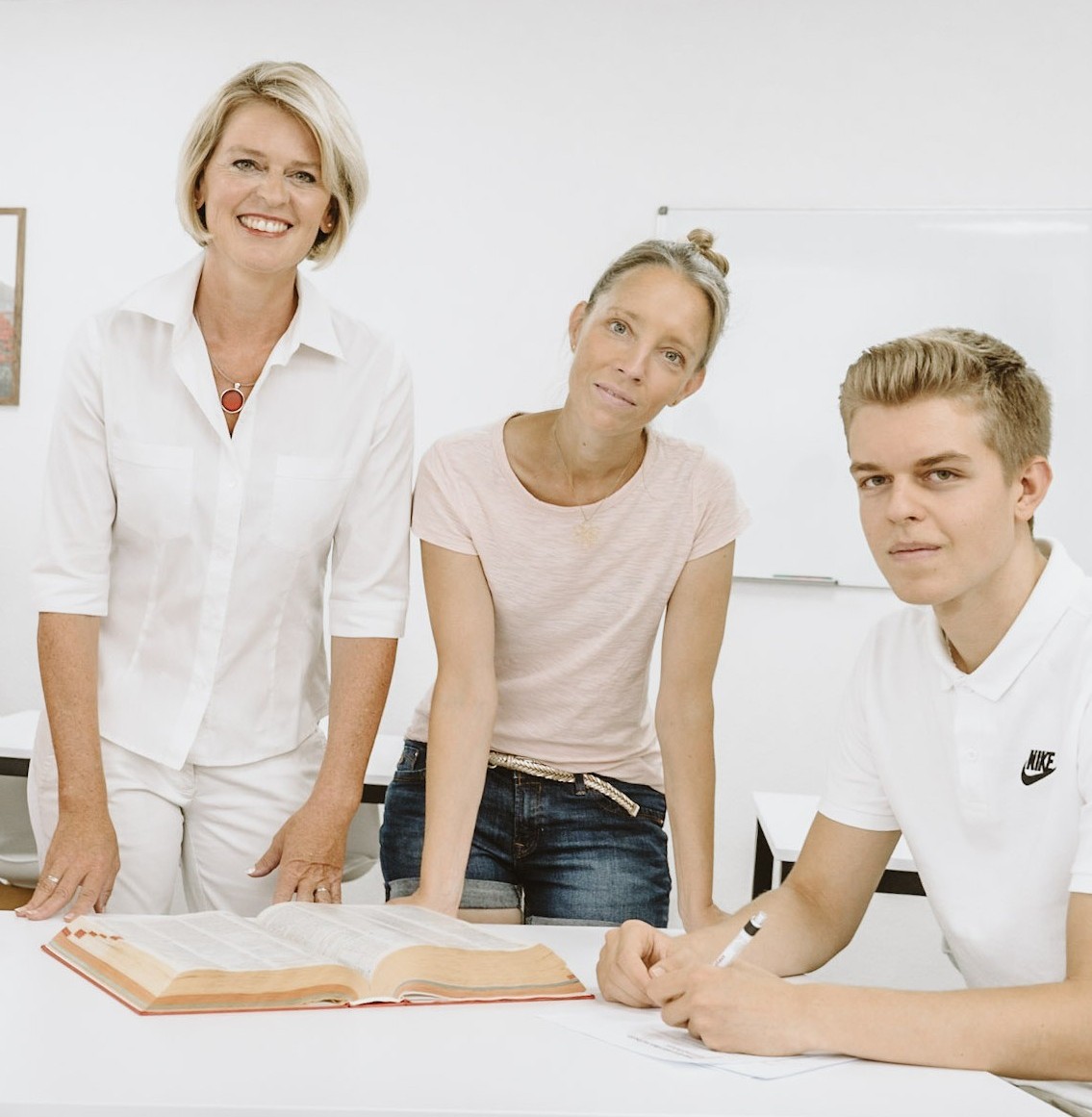 Gruppe-Buch-in-die-Kamera3 Drei Personen stehen in einem hellen Unterrichtsraum an einem Tisch mit einem aufgeschlagenen Fachbuch und arbeiten gemeinsam an Inhalten.